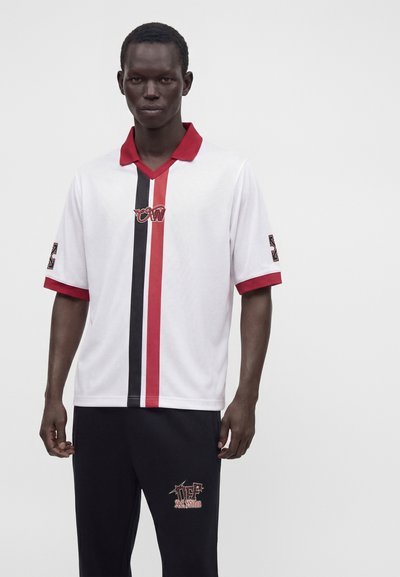 Homme portant une chemise blanche avec un col rouge et des rayures verticales rouges et noires, associée à un pantalon noir avec un logo rouge et blanc.