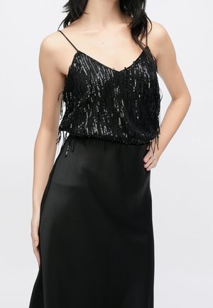 Femme portant un haut noir sans manches avec des sequins brillants et des bretelles fines, associé à une jupe noire lisse, main sur la hanche.
