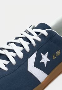 Zapatillas de ante azul con cordones blancos, que presentan un diseño de estrella blanca y la inscripción dorada "ALL STAR", y una suela de goma.