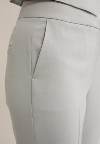 Pantalones ajustados en gris claro hechos de una tela suave, con detalle de pliegue y bolsillos laterales discretos. Tienen un corte entallado.