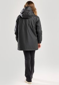 Vesimittainen musta takki hupparin kanssa, joustavat hihansuut ja hieman pidempi leikkaus, näkyy takaa, yhdistetty tummiin housuihin ja vaaleisiin kenkiin.