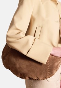 Borsa in suede marrone con bordi cuciti. Stretta nella parte superiore e più ampia in quella inferiore. Abbinata a una giacca in tessuto color crema.