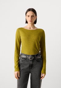 Langermet topp i olivengrønn, laget av teksturert stoff. Kombinert med høyhåndterte svarte jeans og et naglet svart belte.