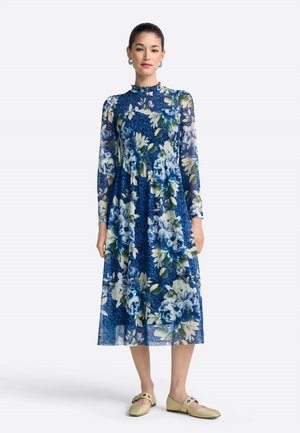 Kleid mit Blumenmuster, blauer Hintergrund mit großen weißen und grünen Blumen, lange Ärmel und eine geraffte Taille. Kombiniert mit hellgrünen Schuhen.