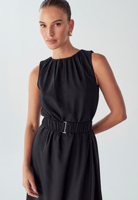 Donna con capelli all'indietro, che indossa orecchini a cerchio dorati e un vestito nero senza maniche con cintura su uno sfondo chiaro.