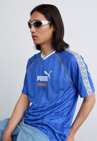 Modrá dres Puma King s vertikálnymi pruhmi, okrúhlym výstrihom a čierno-bielymi vzorovanými detailmi na rukávoch, doplnený o strieborné slnečné okuliare.