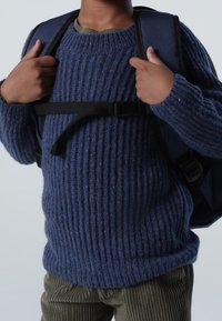 Niebieski sweter dżersejowy z grubą strukturą, połączony z oliwkowymi spodniami sztruksowymi. Czarna plecak z regulowanymi paskami. Dziecięce ręce chwytają za paski.