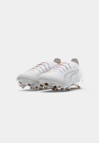 Crampons de football blancs avec une surface texturée, dotés d'accents gris, de coutures orange et d'un maintien en métal au talon. Conçus pour la performance.