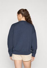 Dunkelblaues Sweatshirt mit Rundhalsausschnitt, lockerer Passform und gerippten Bündchen. Weicher Stoff, minimalistisches Design und ein kleines rundes Detail auf der Rückseite.
