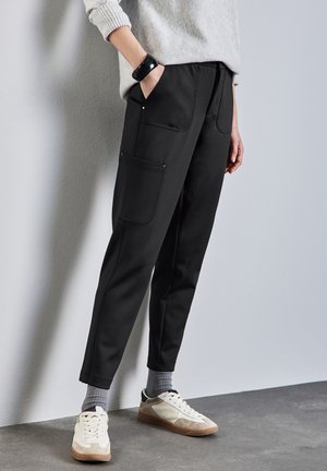 Pantalons noirs cintrés avec poches latérales, fabriqués à partir d'un matériau doux. Portés avec des baskets blanches et des chaussettes grises, sur un fond neutre.