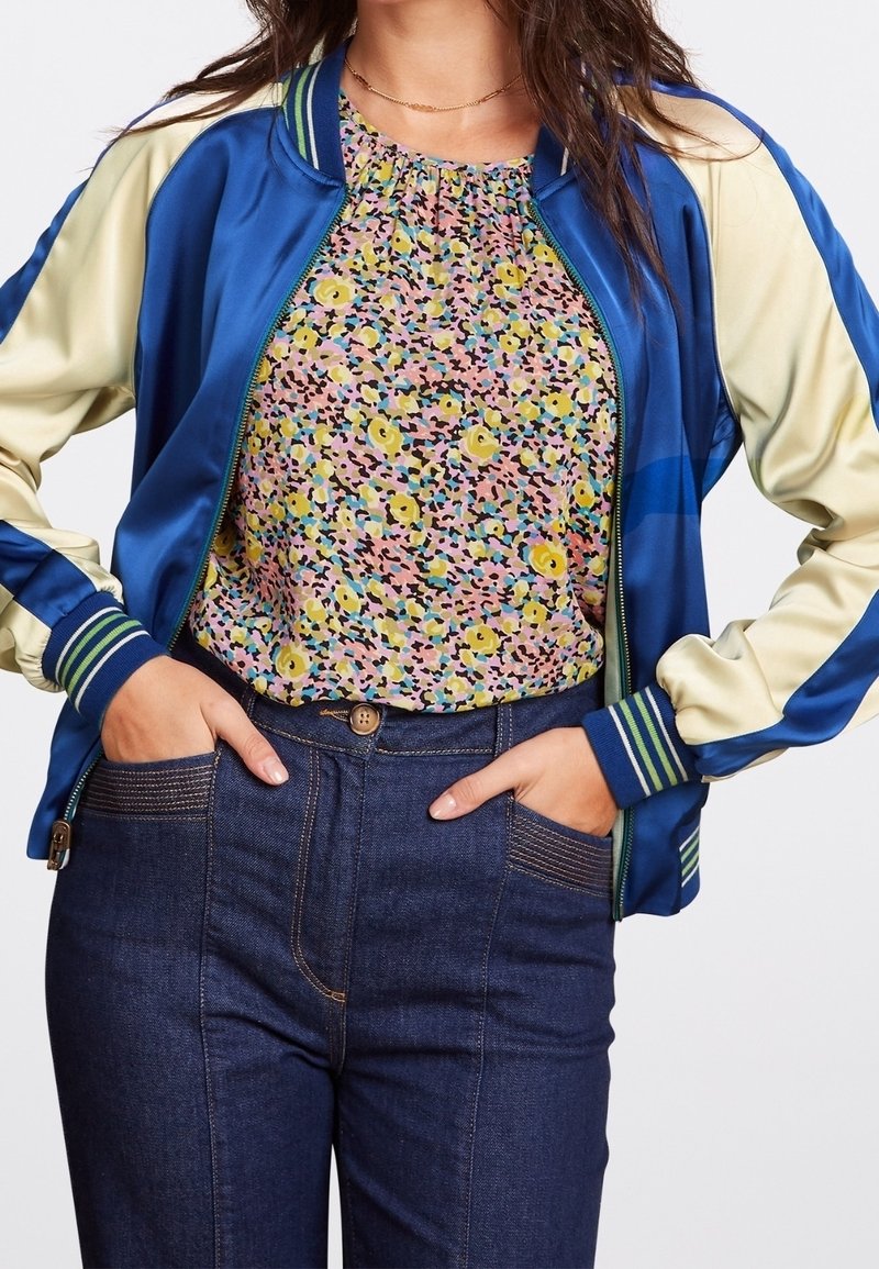 Bomber jacket in satin blu e beige con polsini a righe abbinato a una maglia con motivo floreale, indossato con jeans di denim scuro.