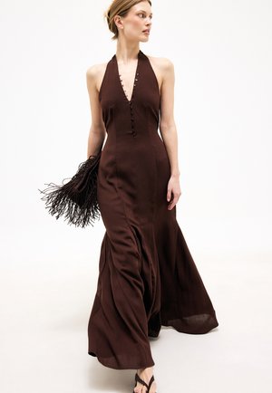 Kobieta w ciemnobrązowej sukience maxi z wiązaniem na szyi, trzymająca torebkę z frędzlami, idąca i patrząca na bok na jednolitym białym tle.