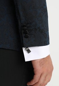 Polso di un uomo con manica di giacca a motivi blu scuro, polsino di camicia bianca, gemello nero e mano rilassata su uno sfondo bianco.