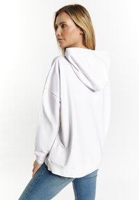 Bílá oversized mikina s kapucí na stažení, s hladkým povrchem a ribbovaným lemem, kombinovaná se světle modrými skinny džíny.