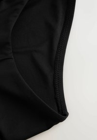 Sort badedragtstof med en glat struktur. Har en buet kant og forstærkede syninger. Nærbillede, der viser materialets detaljer.