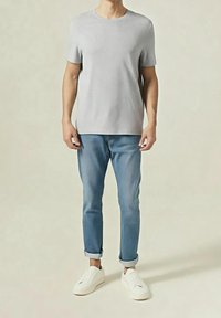 Homme portant un t-shirt à manches courtes gris clair, un jean bleu retroussé et des baskets blanches, debout devant un fond clair uni.