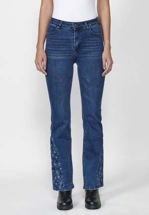 Kvinde iført blå jeans med blomsterbroderi på indersiden af underbenene, kombineret med sorte støvler og en hvid top, stående mod en hvid baggrund.