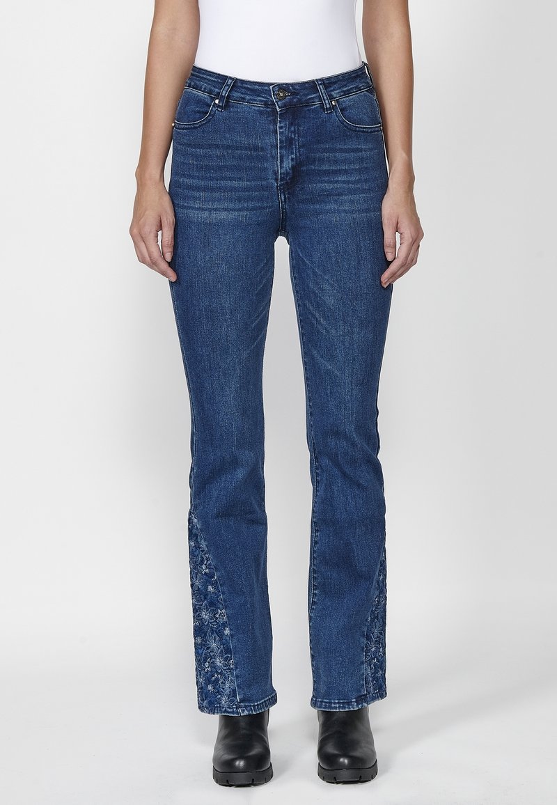 Femme portant un jean bleu avec une broderie florale sur l’intérieur des jambes basses, assorti à des bottes noires et un haut blanc, debout devant un fond blanc.