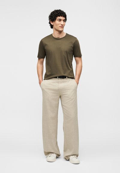 Άνδρας με σγουρά μαλλιά, φοράει ελιάς πράσινο μπλουζάκι, μπεζ παντελόνια με φαρδιά μπατζάκια και μαύρη ζώνη, λευκά αθλητικά παπούτσια, στέκεται με τα χέρια στις τσέπες.