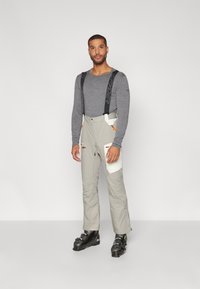 Camisa de manga larga gris debajo de pantalones de esquí gris claro con tirantes negros ajustables, que presentan acentos blancos y cremalleras naranjas, combinados con botas de esquí negras.