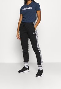 Granatowa koszulka bawełniana z białym logo "adidas", w połączeniu z czarnymi dresami z białymi paskami po bokach oraz czarnymi butami sportowymi.