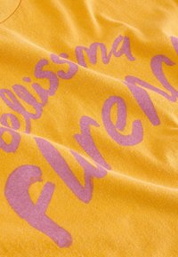 La tela de algodón amarilla presenta un texto en morado claro que dice 'bellissima Firenze'. El material tiene una superficie suave y ligeramente texturizada.