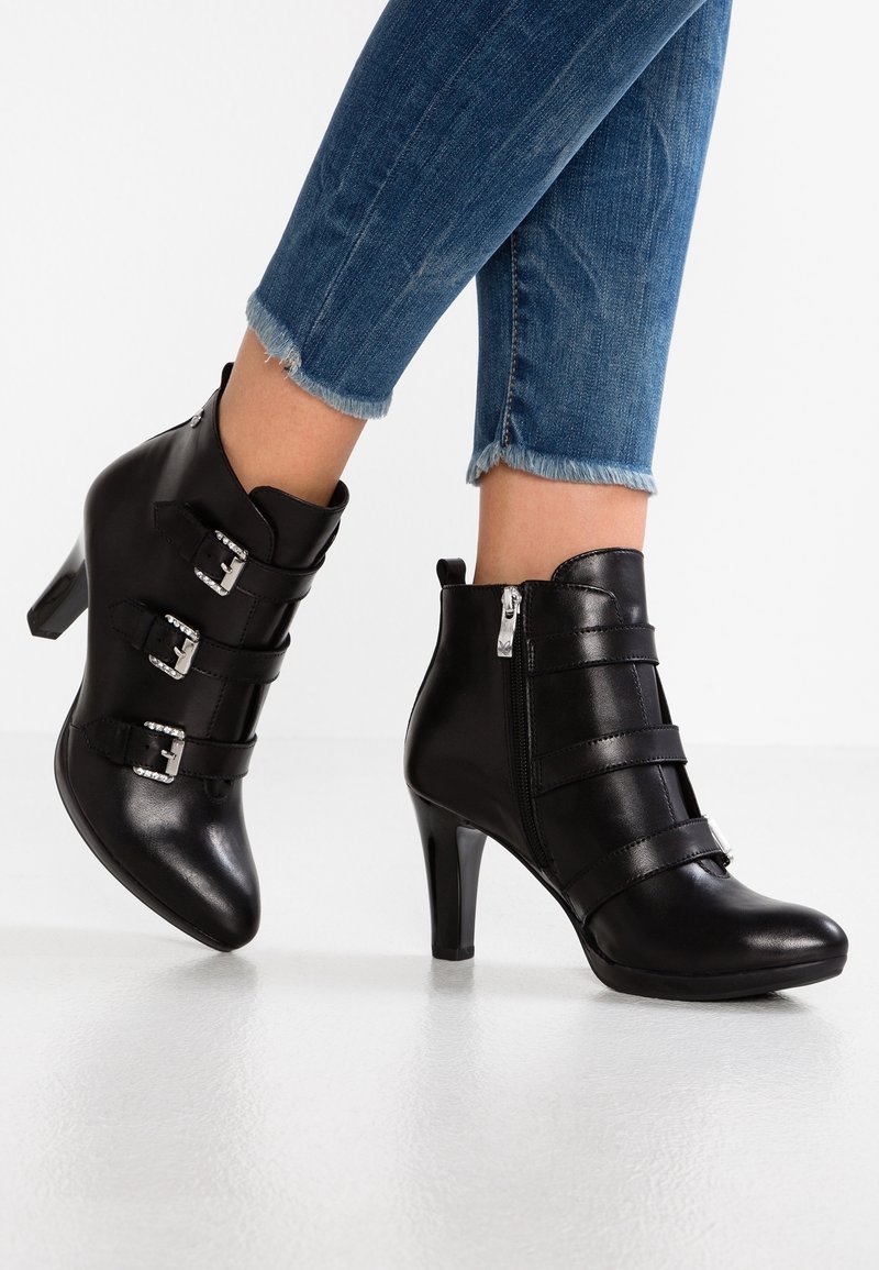 Bottines en cuir noir avec trois boucles décoratives, fermeture éclair latérale et talons hauts, portées avec un jean bleu à ourlet effiloché.