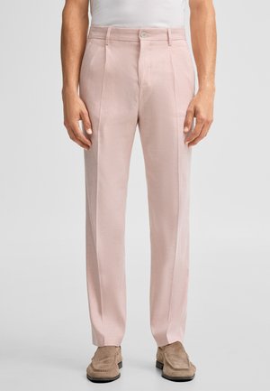 Mann trägt hellrosa maßgeschneiderte Hose und beigen Wildleder-Loafer, steht vor einem schlichten hellen Hintergrund.