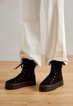 Bottes montantes noires à plateforme avec semelles épaisses en caoutchouc portées avec un pantalon crème à jambes larges sur un sol en bois.