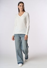 Top bianco a maniche lunghe con scollatura a V, caratterizzato da un piccolo logo ricamato. Abbinato a jeans azzurri a gamba larga e sneakers bianche.