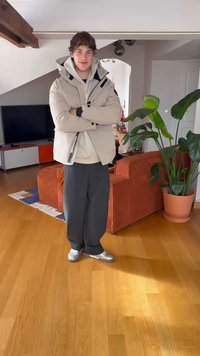 Jachetă puf gri deschis, pantaloni închiși și adidași argintii. Setare interioară cu o plantă și o canapea cu dungi portocalii în fundal.