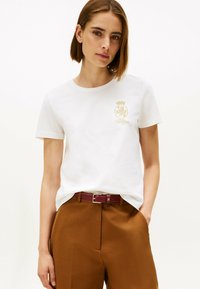 Witte katoenen T-shirt met gouden borduursel van een leeuw en kroon logo, gecombineerd met een bruine broek en een bordeauxrode riem.