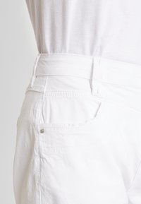 Shorts en coton blancs avec une texture lisse, dotés d'une taille haute, d'une poche latérale et d'un accent métallique sur la poche arrière.