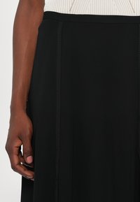 Melnā svārku ar gludu tekstūru, ar plašu jostas daļu un vertikālo šuvju detaļu. Audums brīvi karājas no jostas.