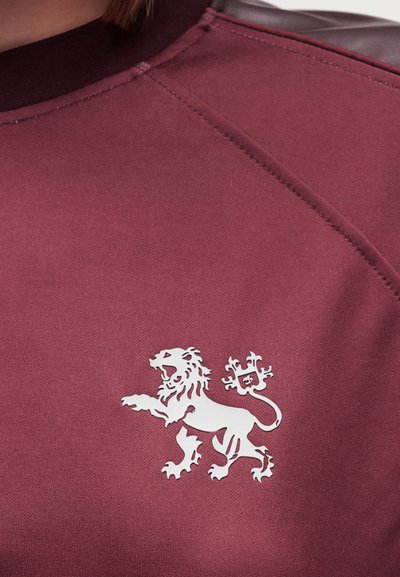 Sweatshirt bordeaux avec un emblème de lion blanc sur le côté gauche de la poitrine, avec des détails de couture distincts et un tissu texturé.
