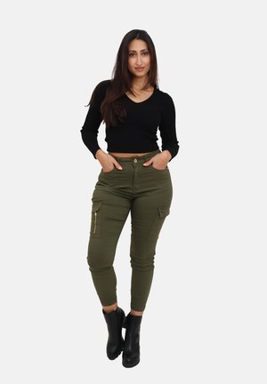 Schwarzes geripptes Crop-Top, kombiniert mit olivgrünen Cargo-Hosen, die mit Reißverschlusstaschen und einer schmal zulaufenden Knöchellänge ausgestattet sind. Das Modell trägt schwarze Knöchelstiefel.