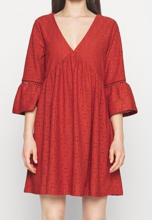 Kvinde iført en rød øjlehulsstofkjole med dyb V-hals og udslåede trekvart ærmer mod en ensfarvet lys baggrund.