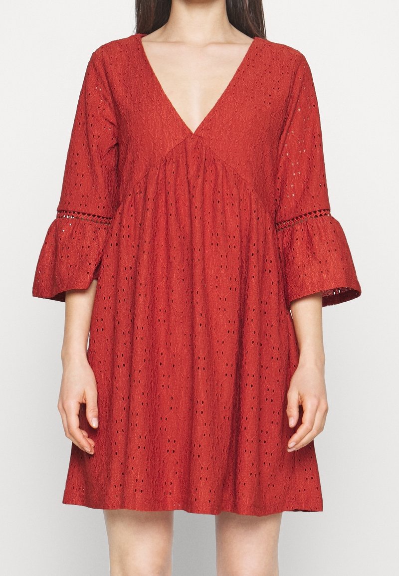 Femme portant une robe rouge en tissu œillet avec un décolleté en V profond et des manches trois-quarts évasées sur un fond clair uni.