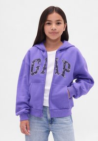 Rapaz com cabelo longo e escuro, vestindo um hoodie roxo da GAP com letras em padrão floral, camisa branca e jeans azul claro, em frente a um fundo simples.