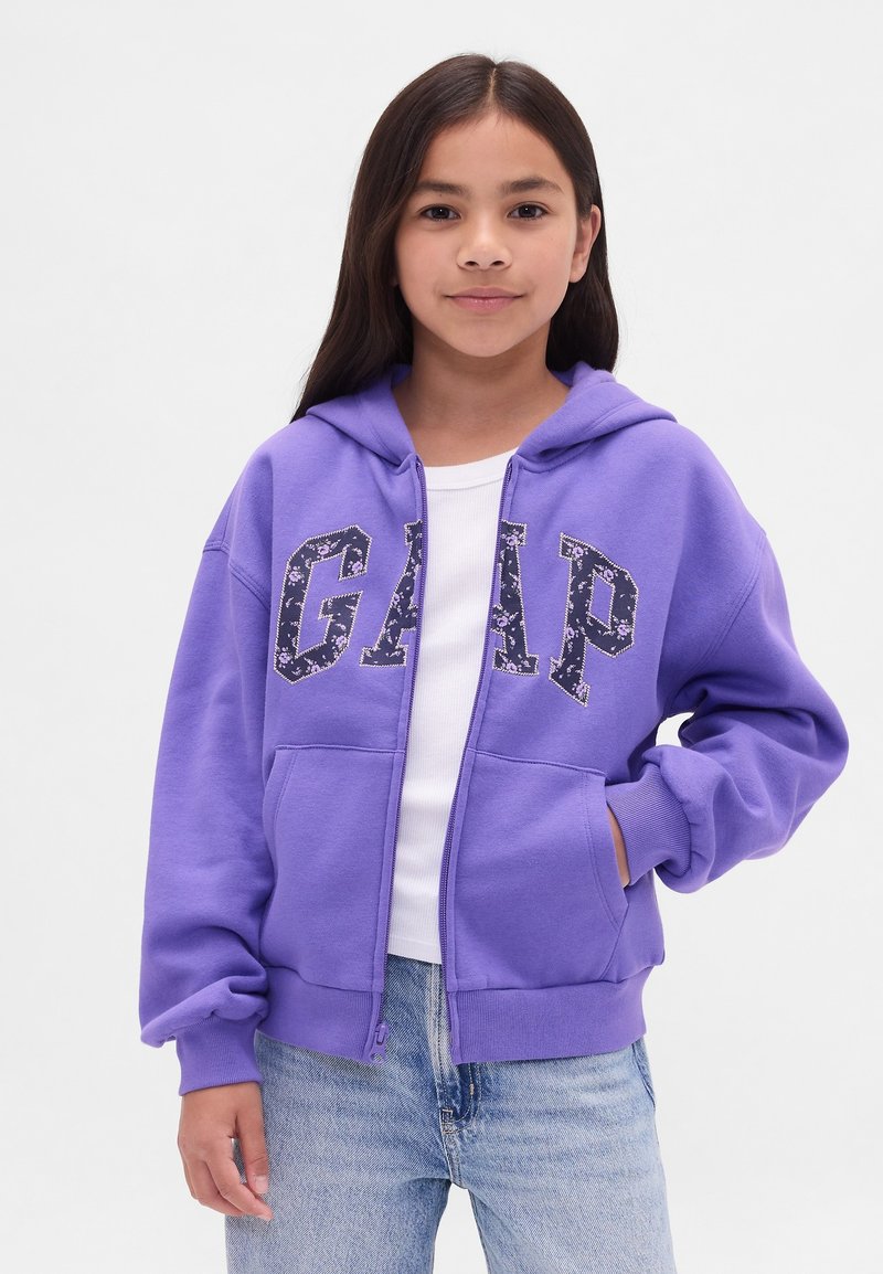Rapaz com cabelo longo e escuro, vestindo um hoodie roxo da GAP com letras em padrão floral, camisa branca e jeans azul claro, em frente a um fundo simples.