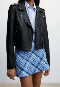 Donna che indossa una giacca in pelle nera da motociclista sopra una camicia bianca, abbinata a una mini gonna a quadri blu con texture.