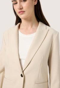 Blazer beige con risvolto strutturato, chiusura con un solo bottone e due tasche frontali. Indossato sopra un top bianco a coste, dalla superficie liscia.