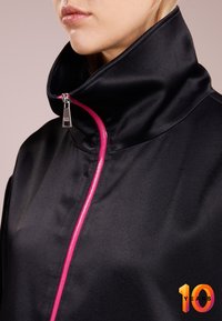 Femme portant une veste noire avec un col haut et une fermeture éclair rose vif partiellement zippée près du cou sur un fond neutre.