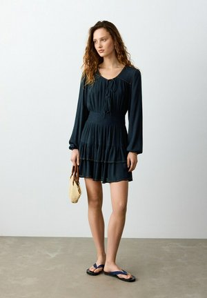 Giovane donna con lunghi capelli ricci che indossa un vestito nero a strati con maniche lunghe, tiene una piccola borsa di paglia e indossa sandali neri.