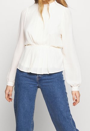 Woman wearing a long-sleeve white blouse with pleated waist detail and blue denim jeans against a plain background.