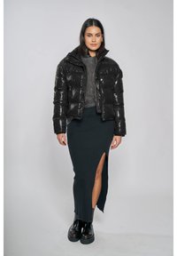 Svart puffjacka med glänsande textur, grå stickad tröja och lång svart kjol med sidospalt, kombinerat med grova svarta stövlar.