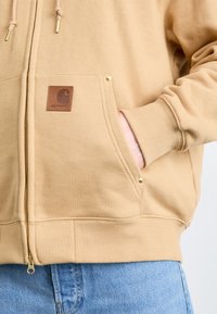 Beige zip-up hoodie with a textured fabric, featuring a leather logo patch, ribbed cuffs, and gold hardware accents on pockets.