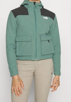 Groene jas met ritssluiting met zwarte schouderstukken en voorflapzakken, gedragen met beige broek, met kleine bloemmotieven tatoeages op de handen.