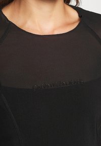 Czarny top Calvin Klein Jeans z gładką teksturą, okrągłym dekoltem i haftowanym logo na środku. Projekt zawiera subtelne szwy.