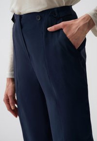 Marineblaue Hose mit glatter Textur, seitlicher Tasche, zwei Knöpfen am Bund und maßgeschneiderter Passform mit dezenten Bügelfalten.